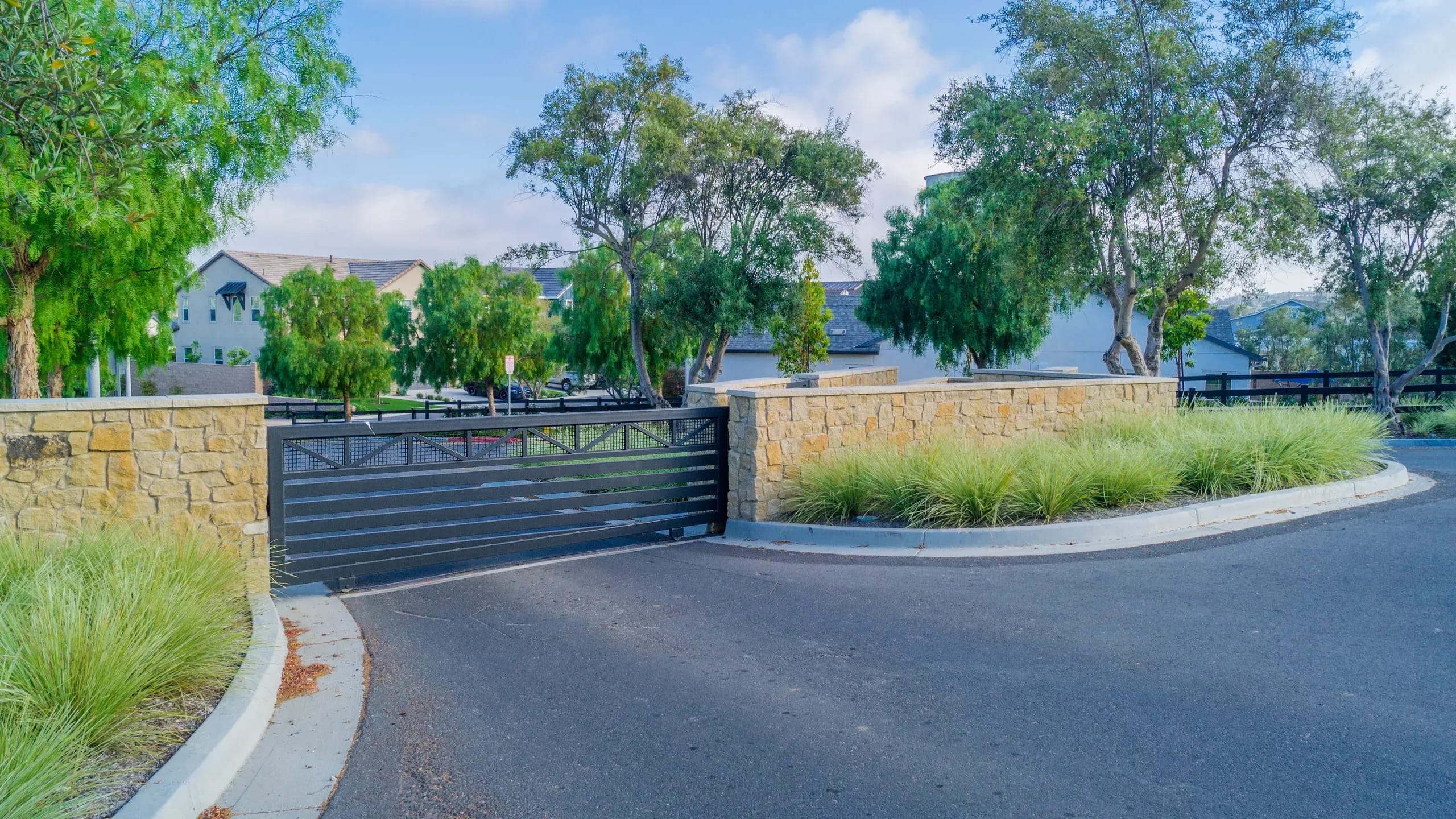 A stone entrance wall and a dark slatted vehicle gate with ornamental grasses and a curved road leading into a gated community under trees.